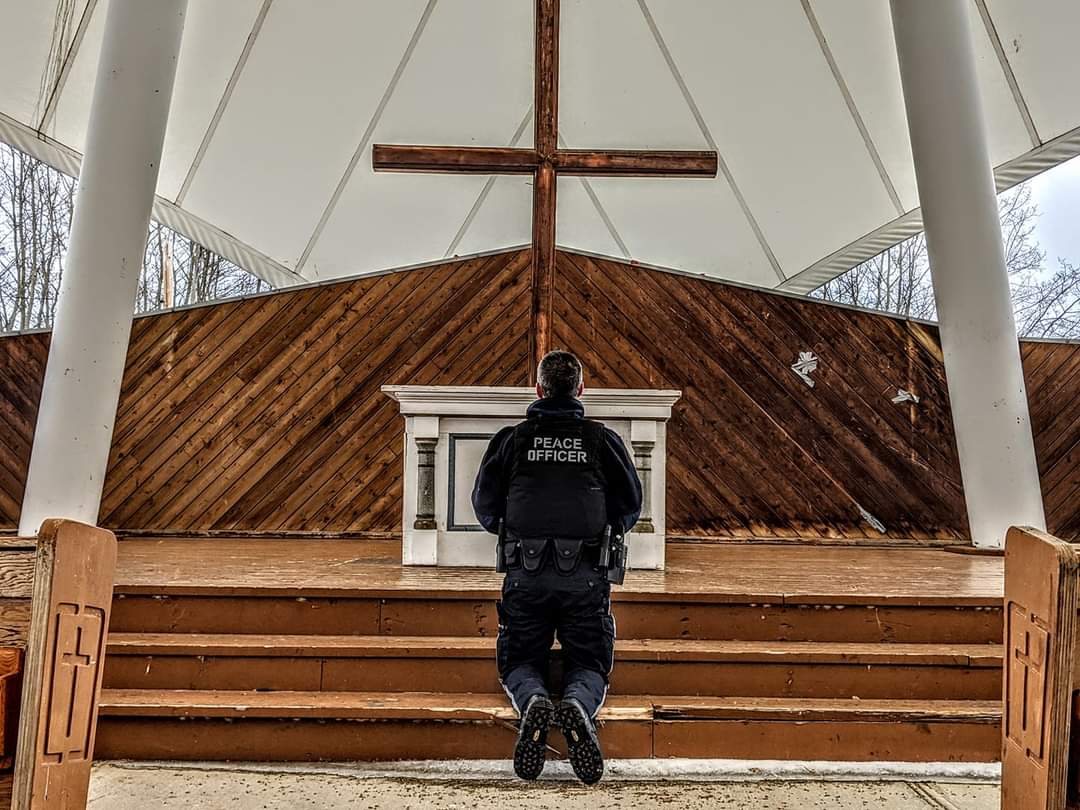 A peace officer kneeling in prayer before a wooden cross at an outdoor chapel