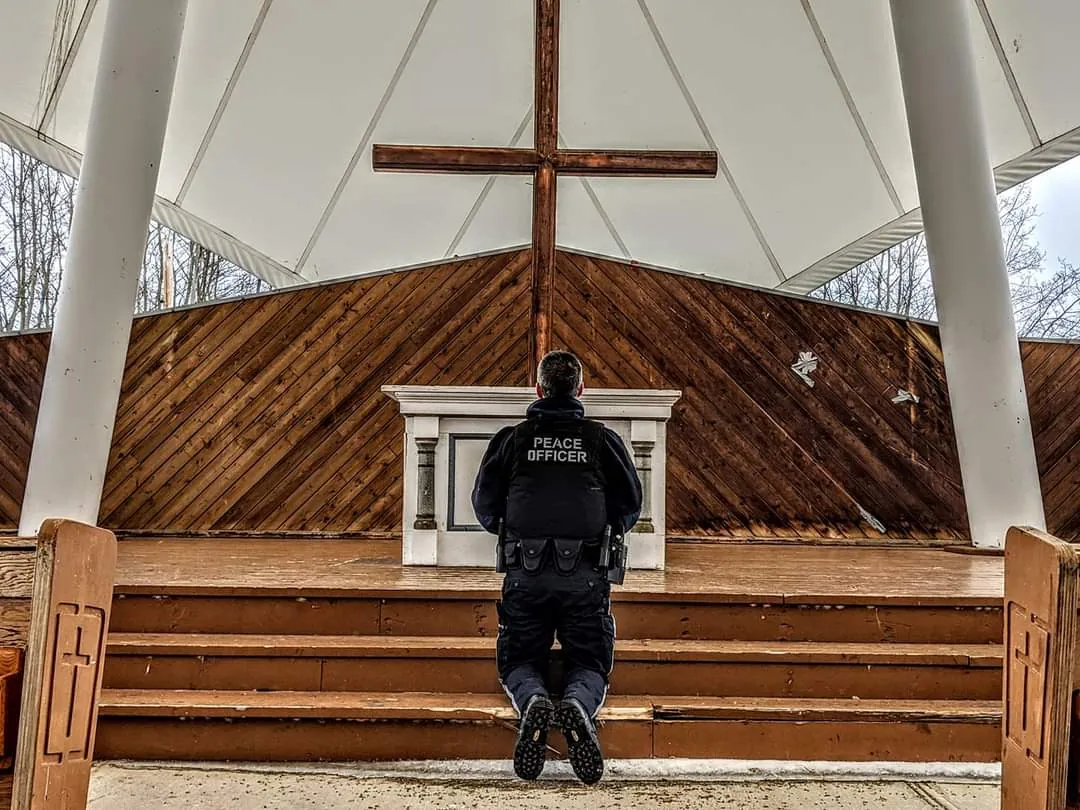 A peace officer kneeling in prayer before a wooden cross at an outdoor chapel